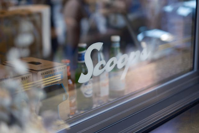 A window with the word Scoops written on it, reflecting a blurred interior scene with bottles and other items visible inside