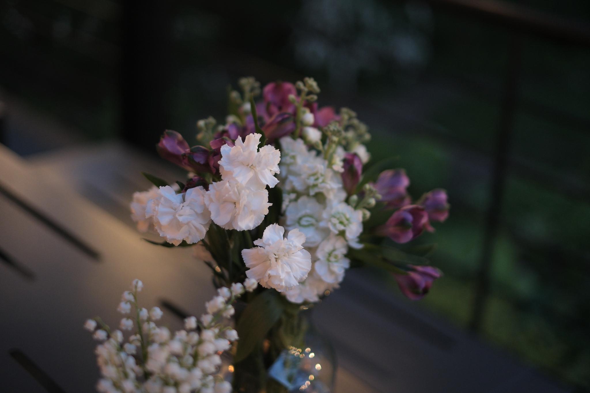 A bouquet of flowers with a mix of white and purple blooms in a glass vase, set on a table with a blurred background suggesting an outdoor setting
