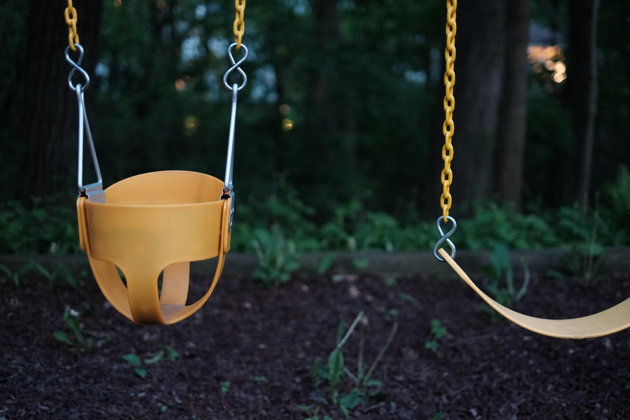 A yellow toddler swing hangs from metal chains against a blurred background of trees and foliage, suggesting a peaceful outdoor setting, possibly a park or a backyard