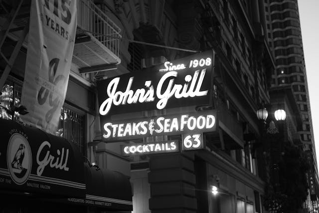 A black and white photograph of a neon sign for John's Grill advertising steaks, seafood, and cocktails, with the establishment date of 1908