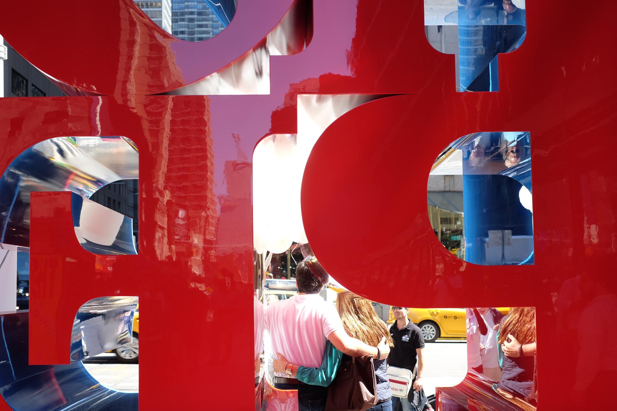 A red sculpture with cut-out letters, through which people and a yellow taxi on a city street are visible