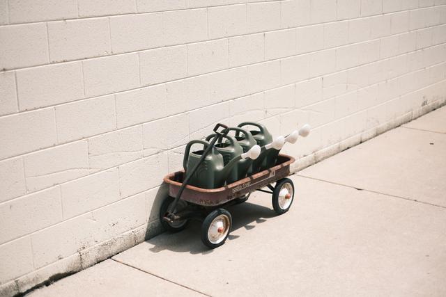 A small red wagon filled with green watering cans is positioned against a white brick wall on a concrete sidewalk