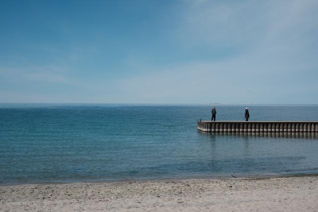 A serene beach with a clear blue sky, calm sea, and two figures standing at the end of a wooden jetty