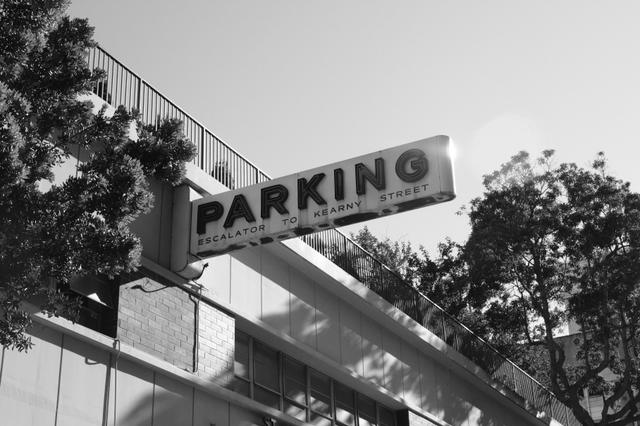 A black and white photograph of a parking sign attached to a building, with trees in the background