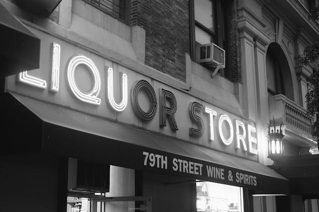 A black and white photograph of a liquor store with a large illuminated sign reading LIQUOR STORE and a smaller sign below it reading 79TH STREET WINE & SPIRITS. The building features classic architectural details and an air conditioning unit in one of the windows
