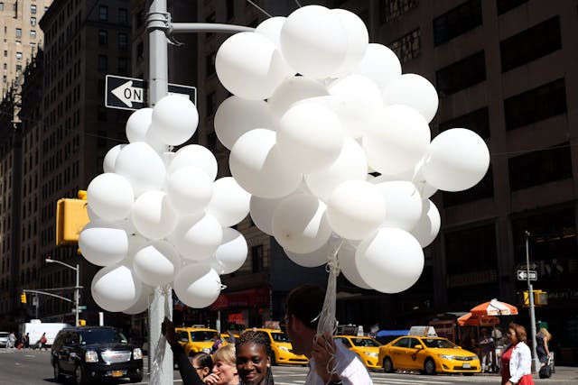 Large clusters of white balloons attached to a street sign in an urban setting, with yellow taxis and tall buildings in the background