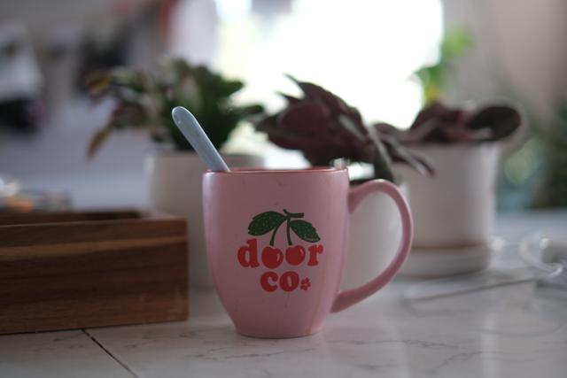 A pink mug with the text deer Co. and a spoon inside, placed on a table with potted plants in the background