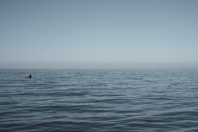A vast expanse of calm ocean water under a clear blue sky, with a single person in the distance