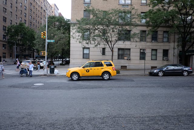A yellow taxi is parked on a city street near a beige building with several windows. Trees and other buildings are visible in the background, and a few pedestrians are walking on the sidewalk