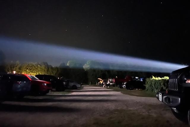 A nighttime scene in a parking area with cars on either side and a bright beam of light stretching across the sky above some trees