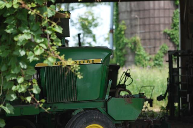A green tractor partially covered by foliage, parked in a barn with a silo visible in the background