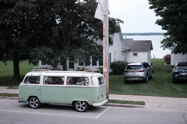 A vintage green and white Volkswagen bus is parked on a residential street with houses and trees in the background, and there's a slight snowy effect overlaying the photo