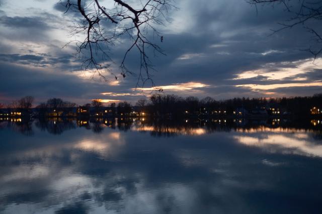 A tranquil evening scene over water, with a reflection of the twilight sky and lights from nearby buildings, accented by silhouette tree branches in the foreground