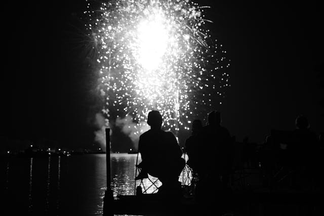 Silhouetted figures sitting on a dock, watching a bright fireworks display over a body of water at night