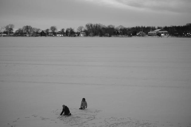 Two people sitting on a frozen lake with a distant shoreline and houses in the background, captured in black and white