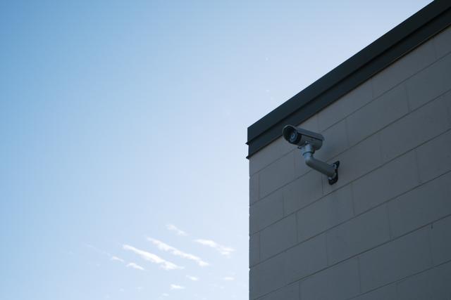A security camera mounted on the corner of a building against a clear blue sky