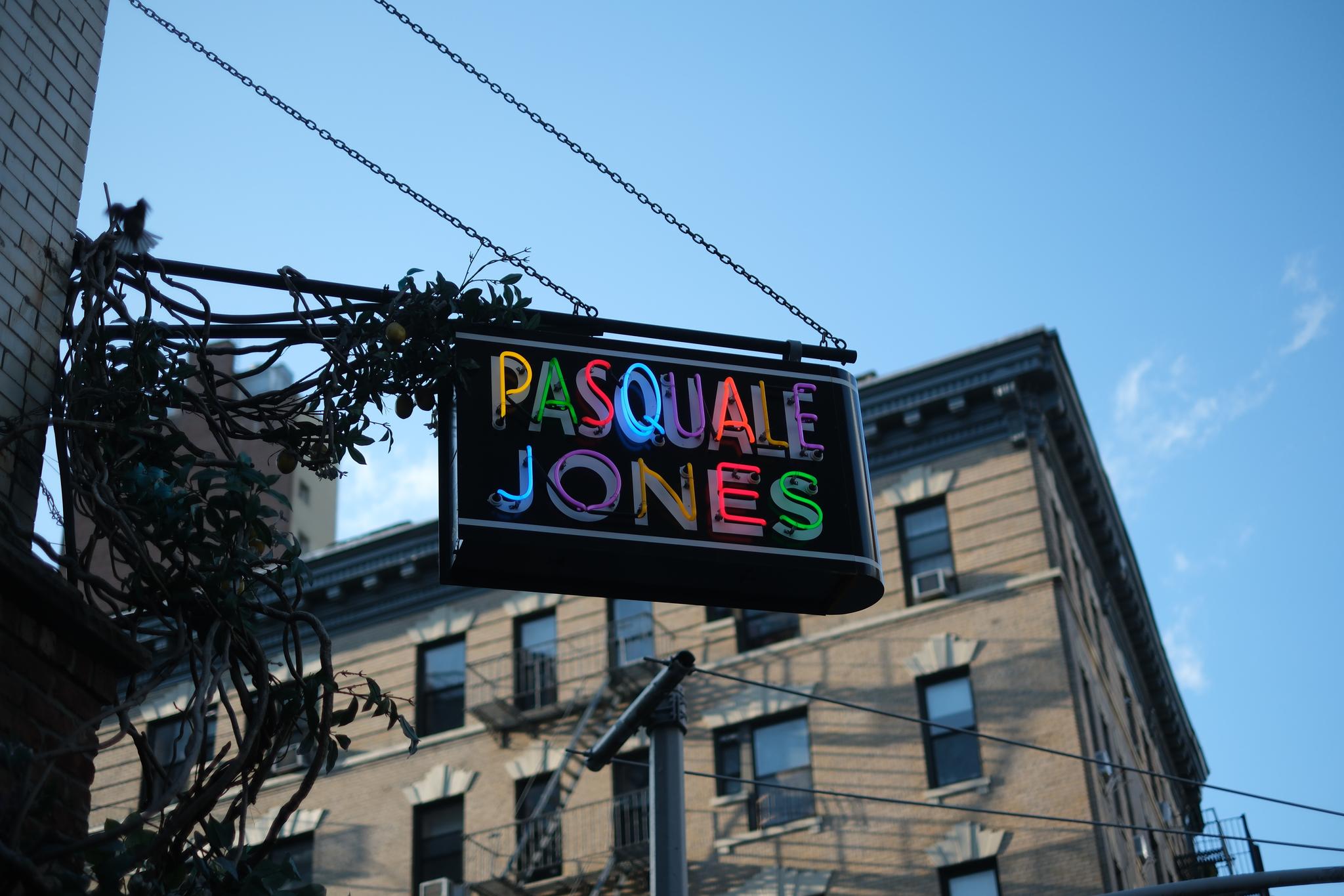 A colorful neon sign reading Pasquale Jones hangs on a metal bracket against a backdrop of a multi-story brick building and a clear blue sky