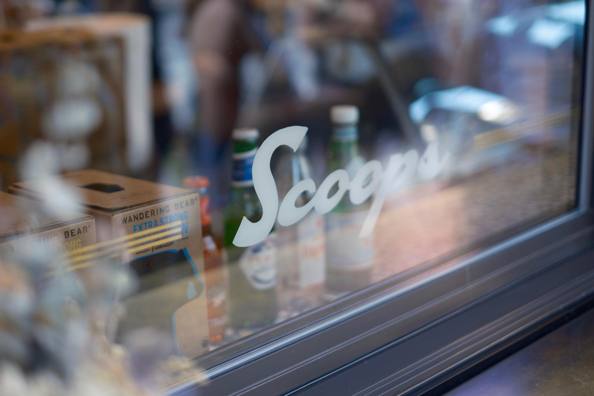 A window with the word Scoops written on it, reflecting a blurred interior scene with bottles and other items visible inside