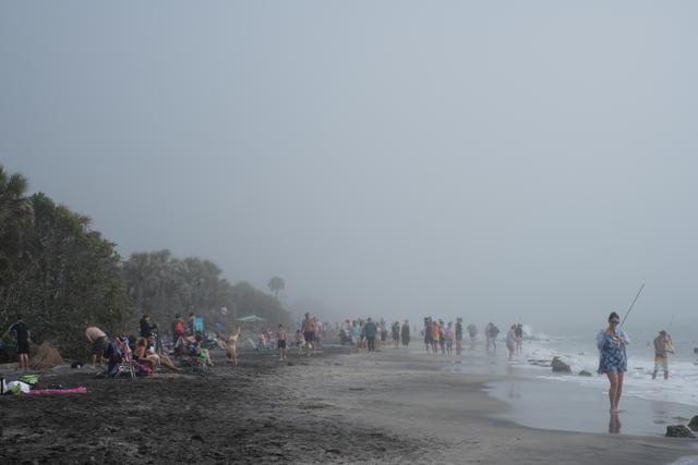A misty beach scene with people scattered along the shore, some in the water, and others lounging or walking on the gray sand