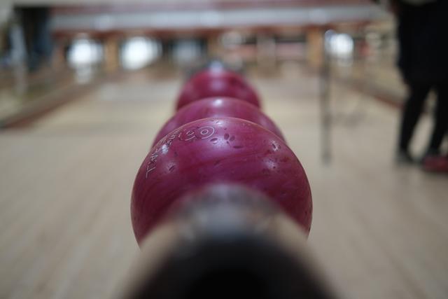 A close-up view of a row of red bowling balls aligned on a rack, seen from the level of the balls, with a bowling lane blurred in the background