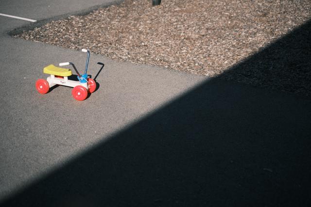 A small tricycle with red wheels and a yellow seat is positioned on a paved surface, partially in shadow, with a gravel area in the background