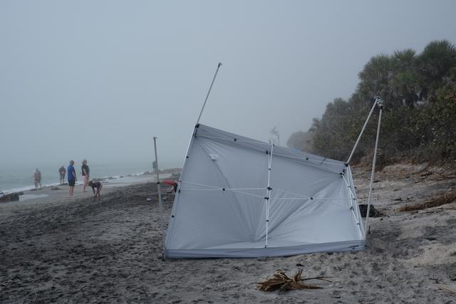 A collapsed canopy on a foggy beach with a few people walking in the distance and trees in the background