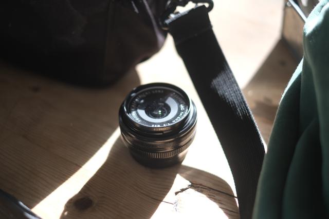 A camera lens placed on a wooden surface, with a strap and a bag nearby, illuminated by natural light