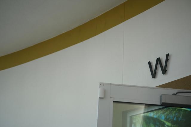 A white wall with a yellow stripe near the ceiling and a black letter W mounted on it, next to a partially open glass door