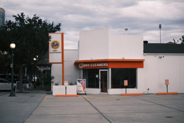 A small, white building with an orange awning and sign, labeled Dry Cleaners, situated in a parking lot with a cloudy sky overhead