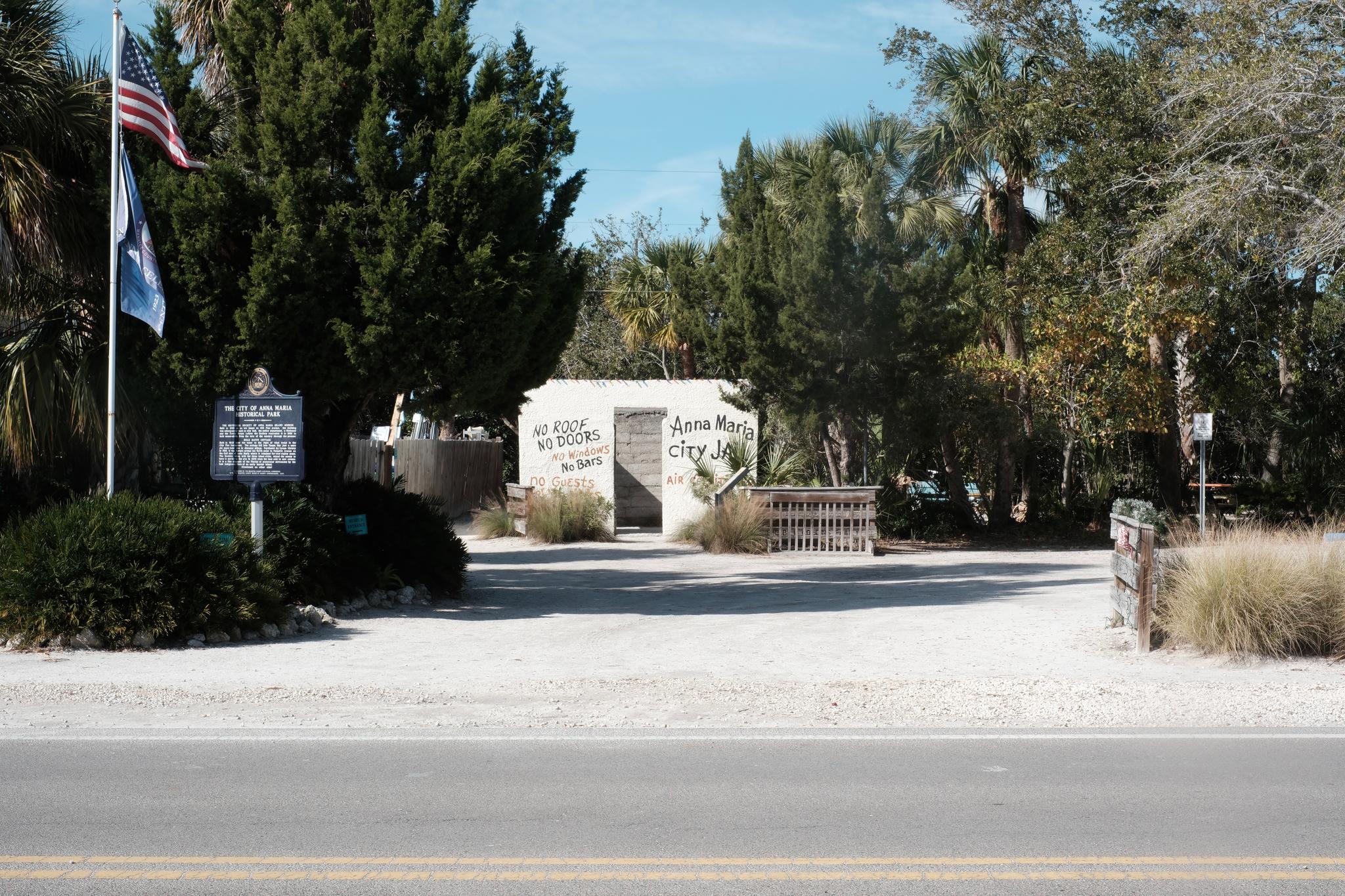 A gated entrance to a park or garden area, with a white building in the background, surrounded by trees and greenery. An American flag and another flag are visible on the left side, along with a sign. The ground is covered with gravel, and a road runs in the foreground