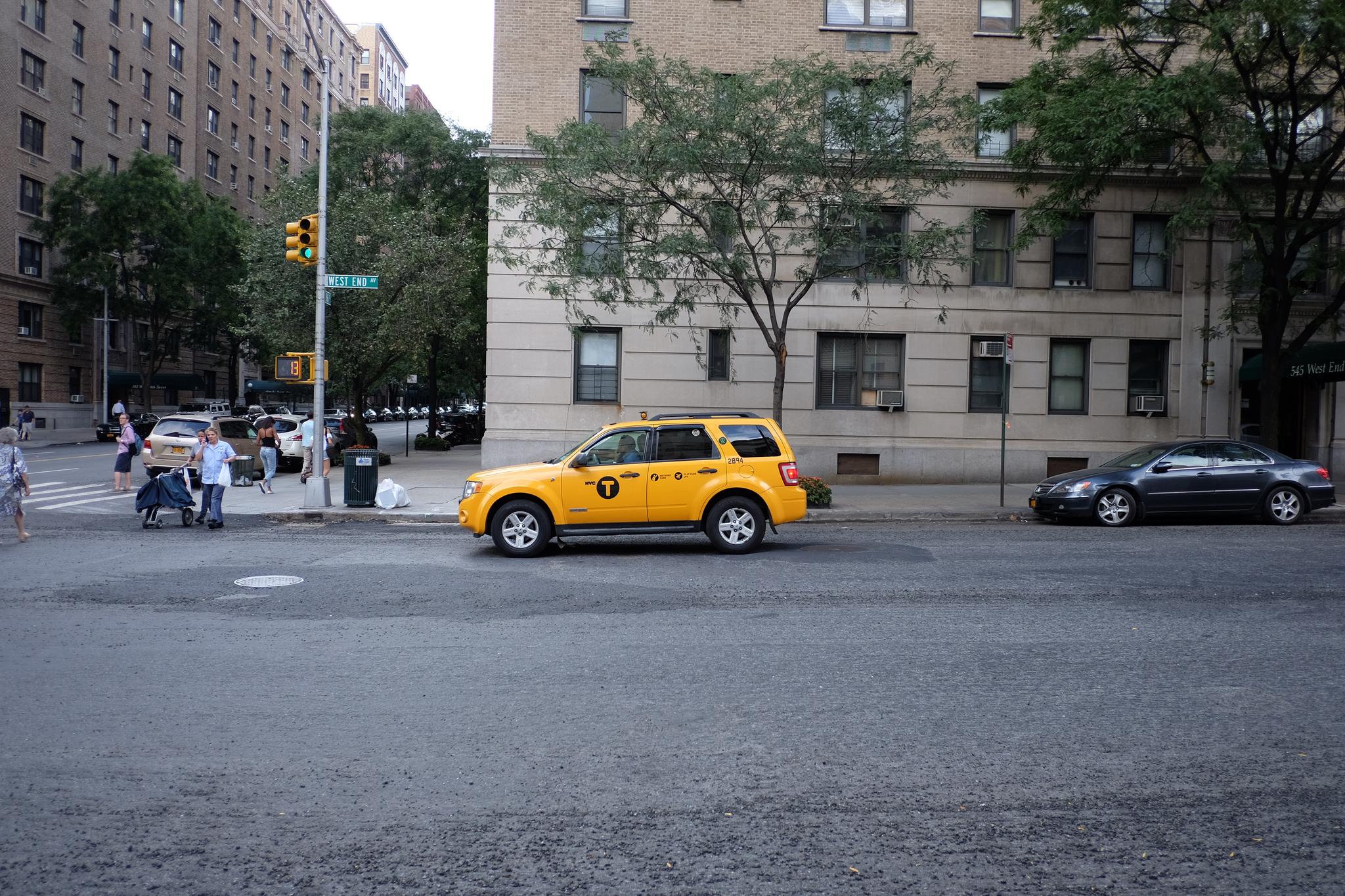 A yellow taxi is parked on a city street near a beige building with several windows. Trees and other buildings are visible in the background, and a few pedestrians are walking on the sidewalk