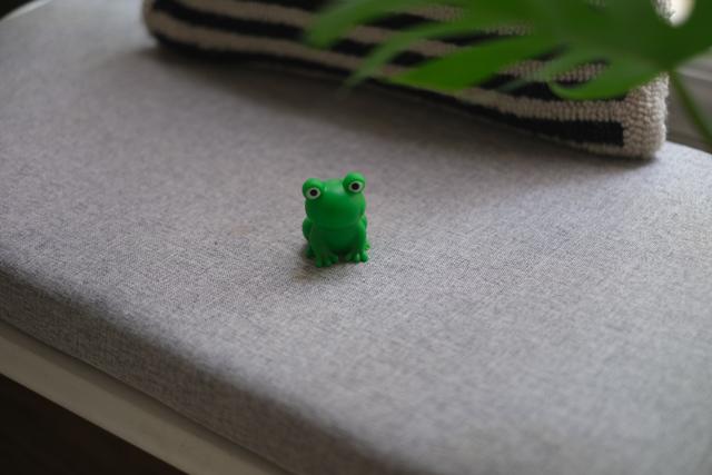A small green frog figurine placed on a gray cushioned surface with a striped pillow and a green plant in the background