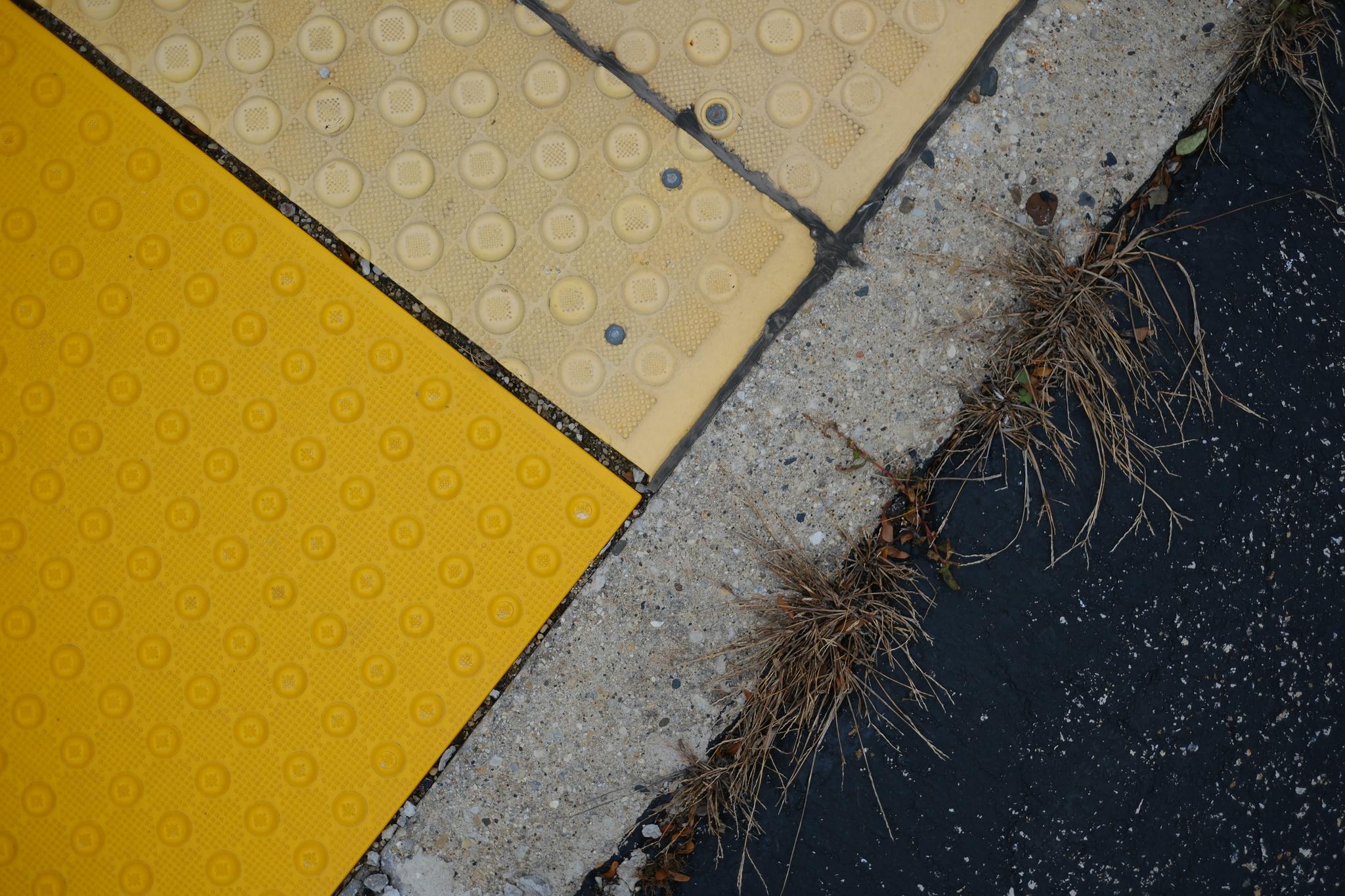 A close-up of a sidewalk corner with yellow tactile paving, adjacent to a concrete curb and asphalt road, with some grass growing in the cracks