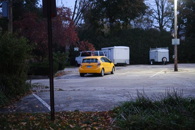 A yellow car is parked in an almost empty parking lot with two white trailers in the background. The scene is set during dusk or early evening, with trees and bushes surrounding the area
