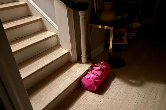 A pink duffle bag rests at the foot of a staircase bathing in a beam of light, with the surrounding area in soft shadows