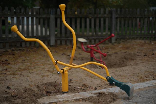 A playground digger toy with yellow handles and a green scoop is set in a sandy area, with a red digger toy in the background and a wooden fence behind them