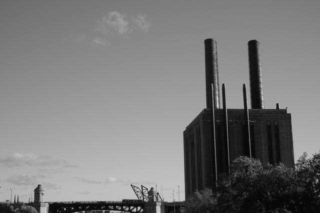 A black and white photograph of an industrial building with tall smokestacks, set against a clear sky with a few clouds. Trees and a bridge are visible in the foreground