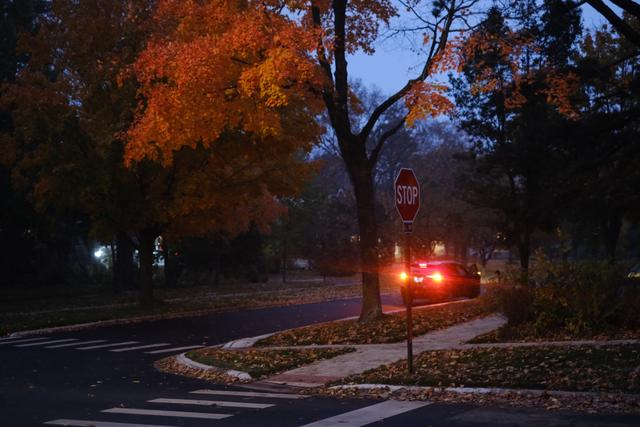 A quiet suburban street at dusk with autumn foliage, a stop sign, and a car with its taillights illuminated
