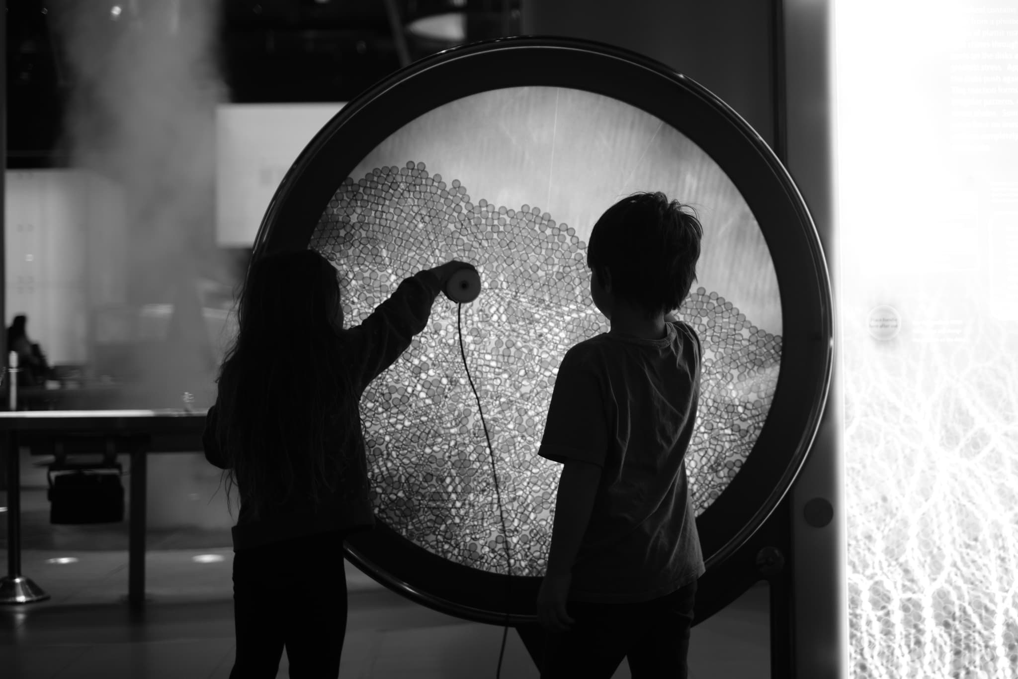 Two children are silhouetted against a large circular interactive display, with one child reaching out to touch it The scene is in black and white, creating a dramatic contrast