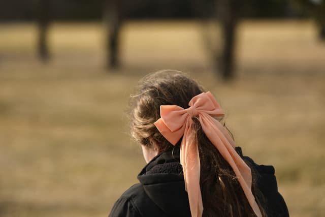 A person with long hair tied in a ponytail with a large peach-colored bow, standing outdoors with a blurred background of trees and grass