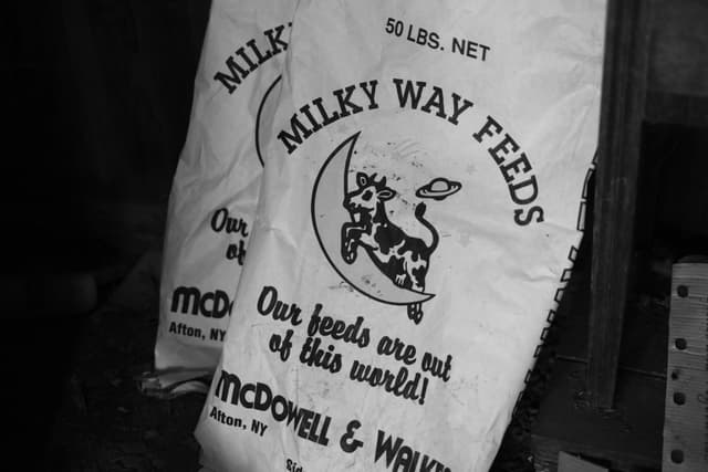 A feed bag labeled MILKY WAY FEEDS with a graphic of a cow jumping over the moon, accompanied by the slogan Our feeds are out of this world! and the names McDowell & Walker