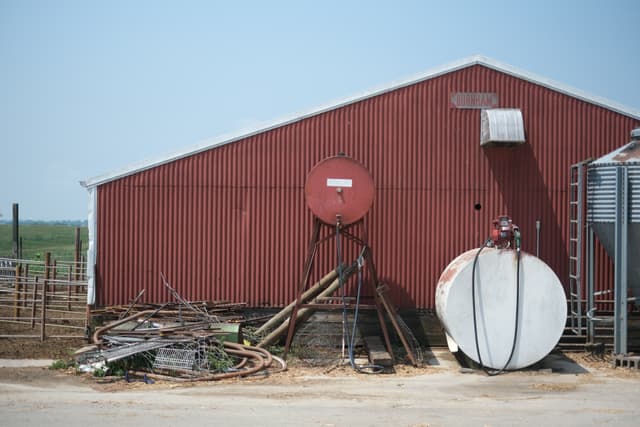 A red barn with a slanted roof, featuring a large round tank and various farming equipment and debris in front of it