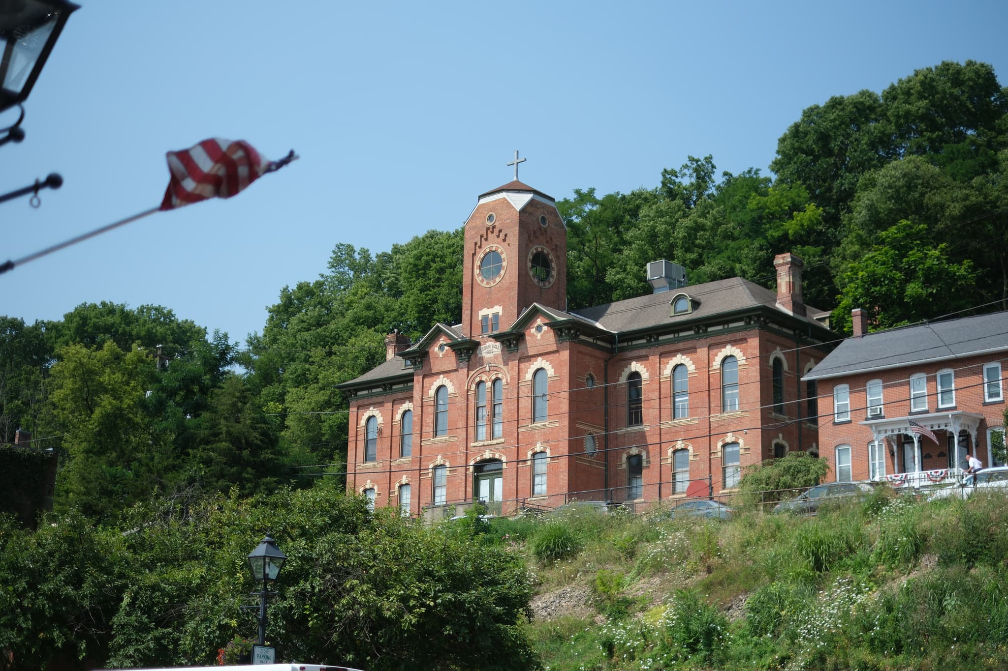 A large, historic brick building with arched windows and a central tower, situated on a hillside with lush greenery in the background. An American flag is visible in the foreground