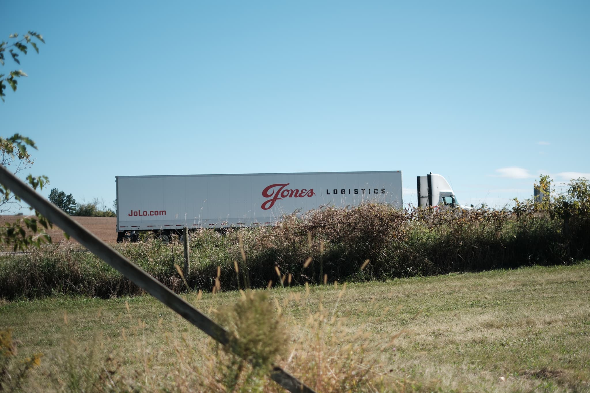 A large white truck trailer with the word Giant and Logistics written on its side, parked in a grassy area with some bushes and trees in the background