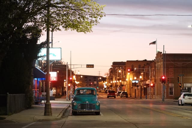 A quiet street scene at dusk with a vintage car parked on the side, illuminated by streetlights and surrounded by buildings