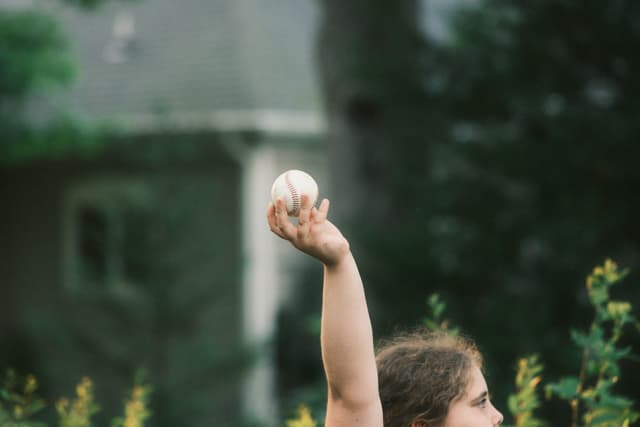 Person outdoors with arm raised holding a small object, with a house and trees blurred in the background