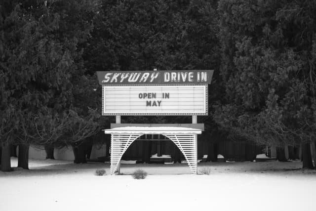A black and white photo of a Skyway Drive In sign surrounded by trees, with the message Open in May