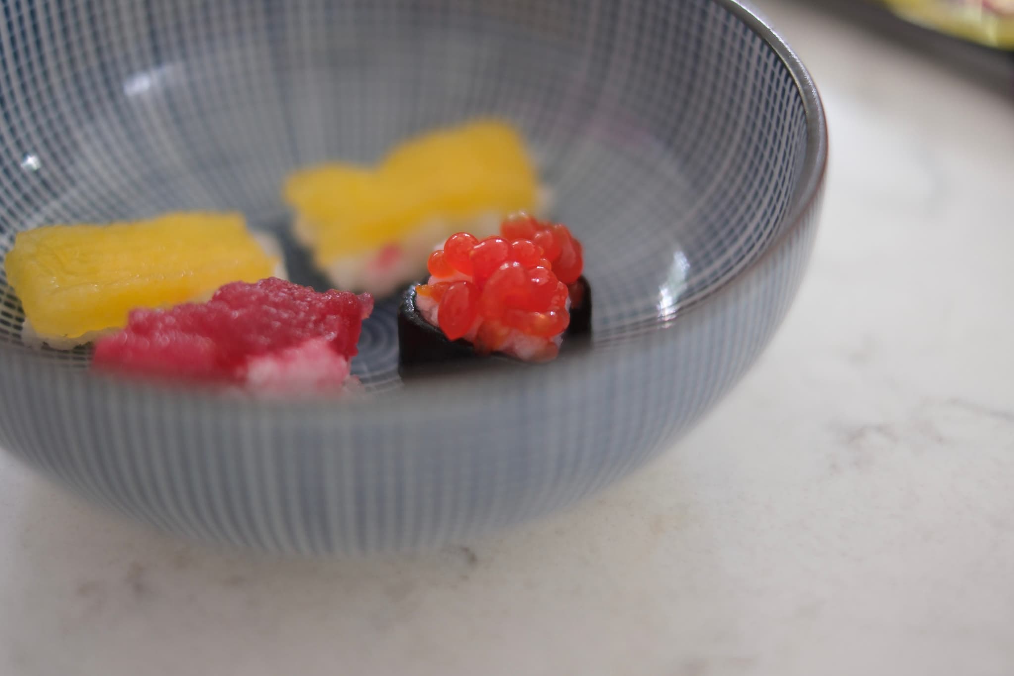 A blue bowl containing pieces of sushi with various toppings, including yellow and red ingredients, placed on a light-colored surface