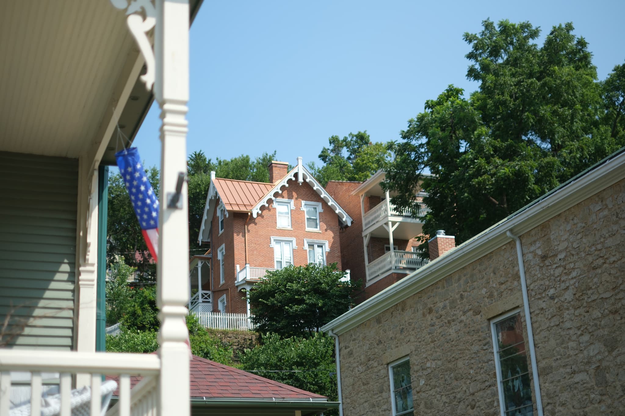 A red brick house with white trim is situated on a hill, surrounded by trees. In the foreground, parts of two other buildings are visible, one with an American flag hanging from a porch