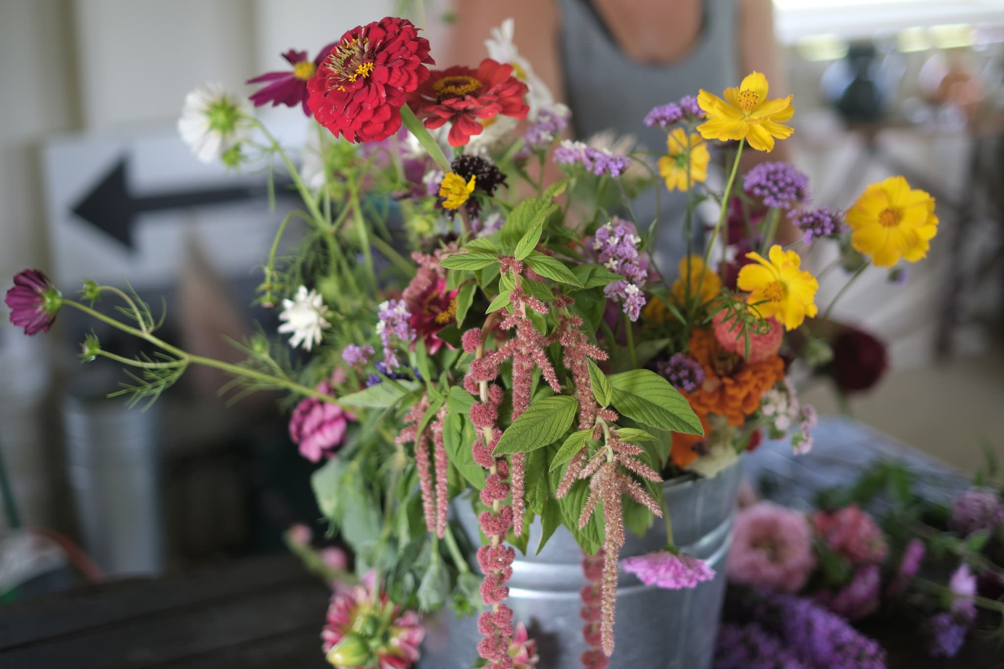 A vibrant bouquet of various flowers in a metal bucket, featuring red, yellow, purple, and white blossoms, with a person in the background